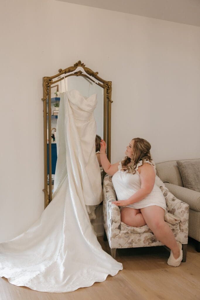 The bride admires her dress that is hanging on the mirror in the dressing room.